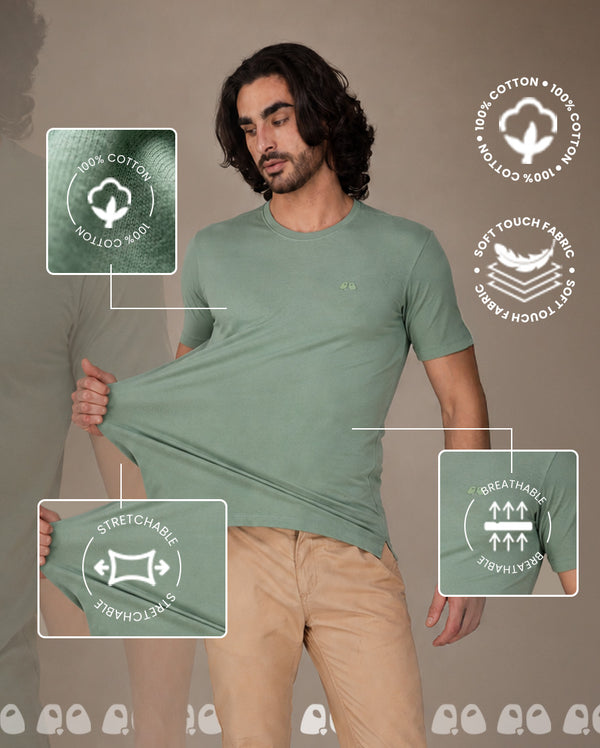 A man wearing a Vandee sage green short-sleeve t-shirt, stretching it to demonstrate its elasticity. 